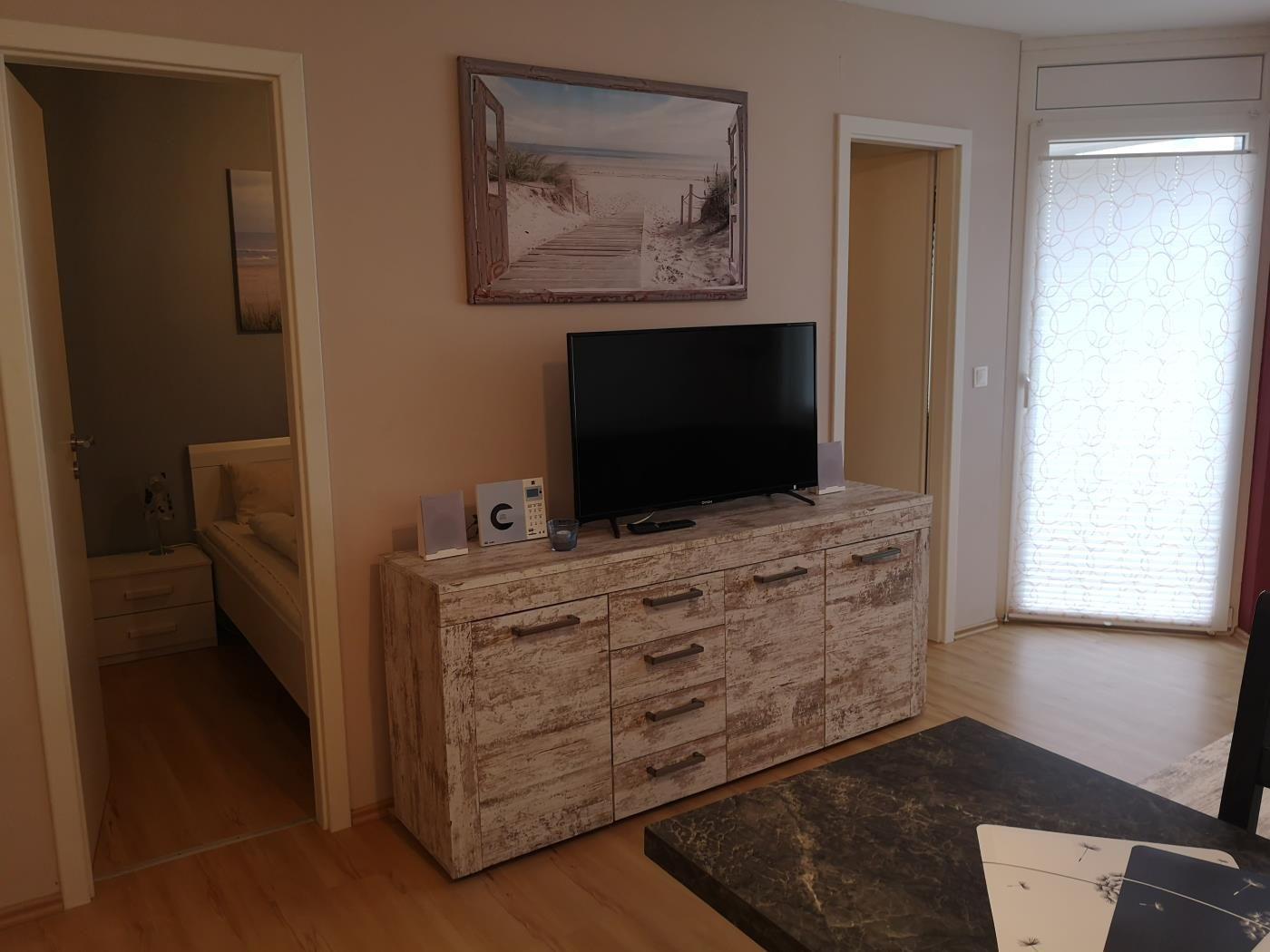 Wohnzimmer mit Fernseher auf Schrank, Blick in Schlafzimmer und Fenster mit Jalousie.