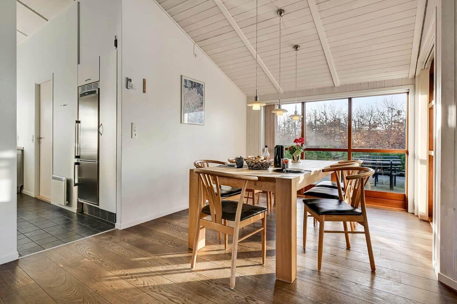 Dining area with wooden table and chairs, view of garden. Kitchen area with refrigerator.