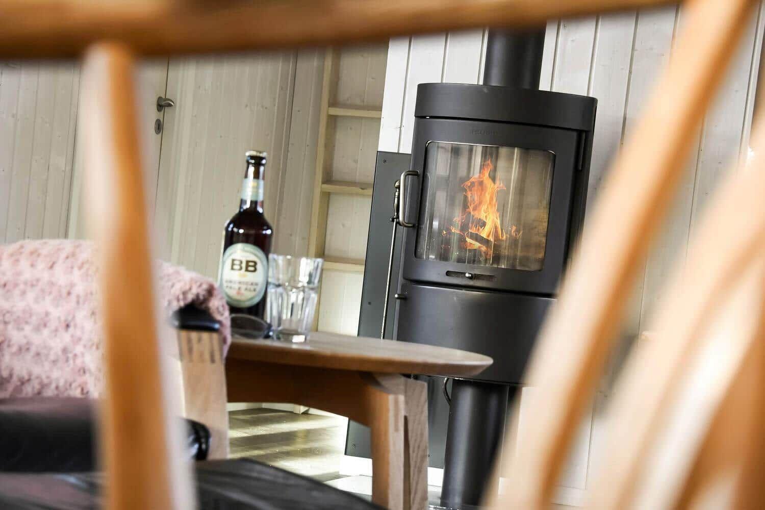 A wood-burning stove stands in a room with wooden walls and a table with a bottle and glasses.