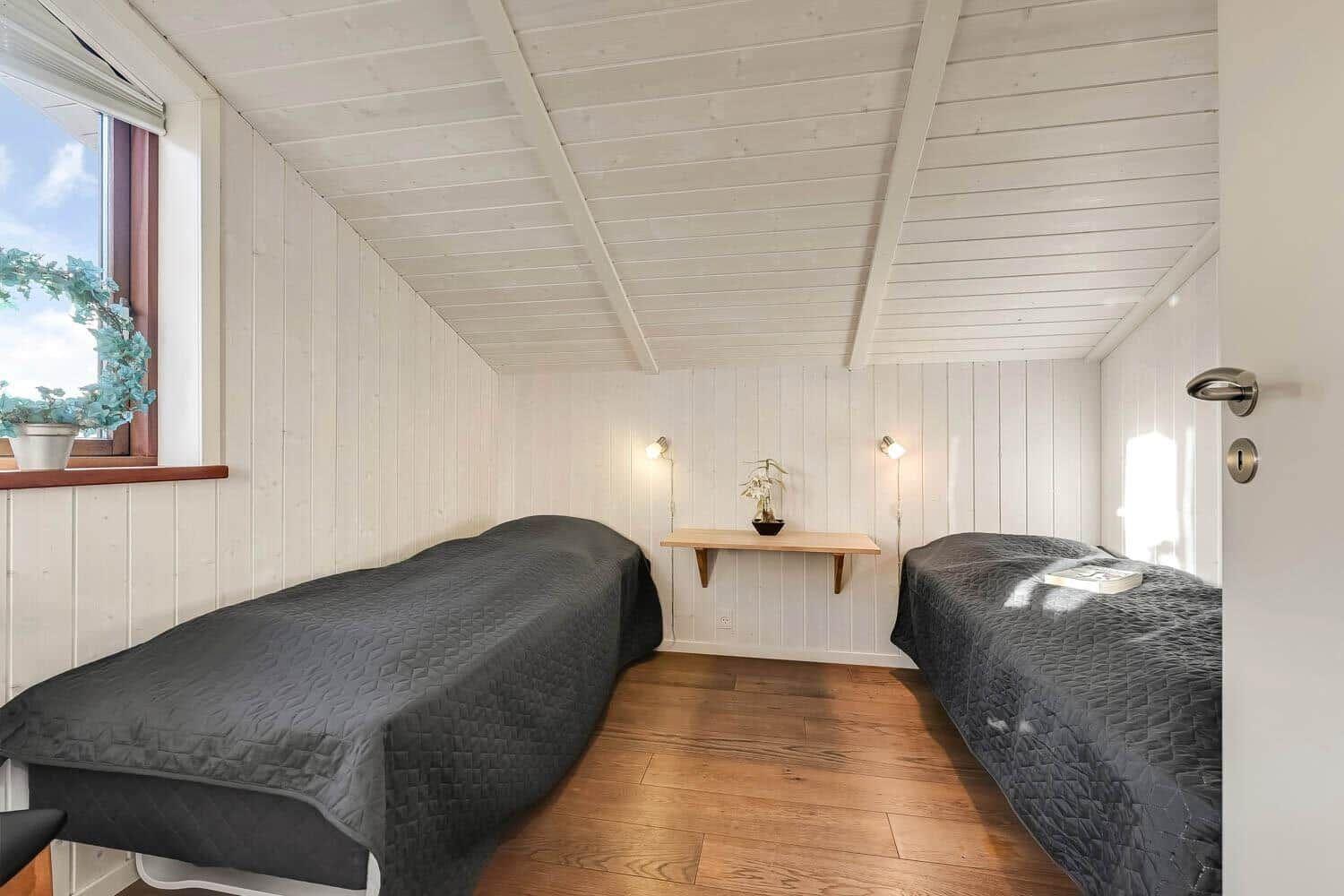 Two beds with dark covers, wooden floor, and white paneled walls with ceiling structure.