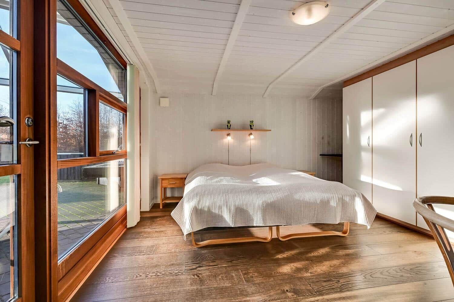 Bedroom with wooden floor, large window, and white bed. A shelf with two lamps hangs on the wall.