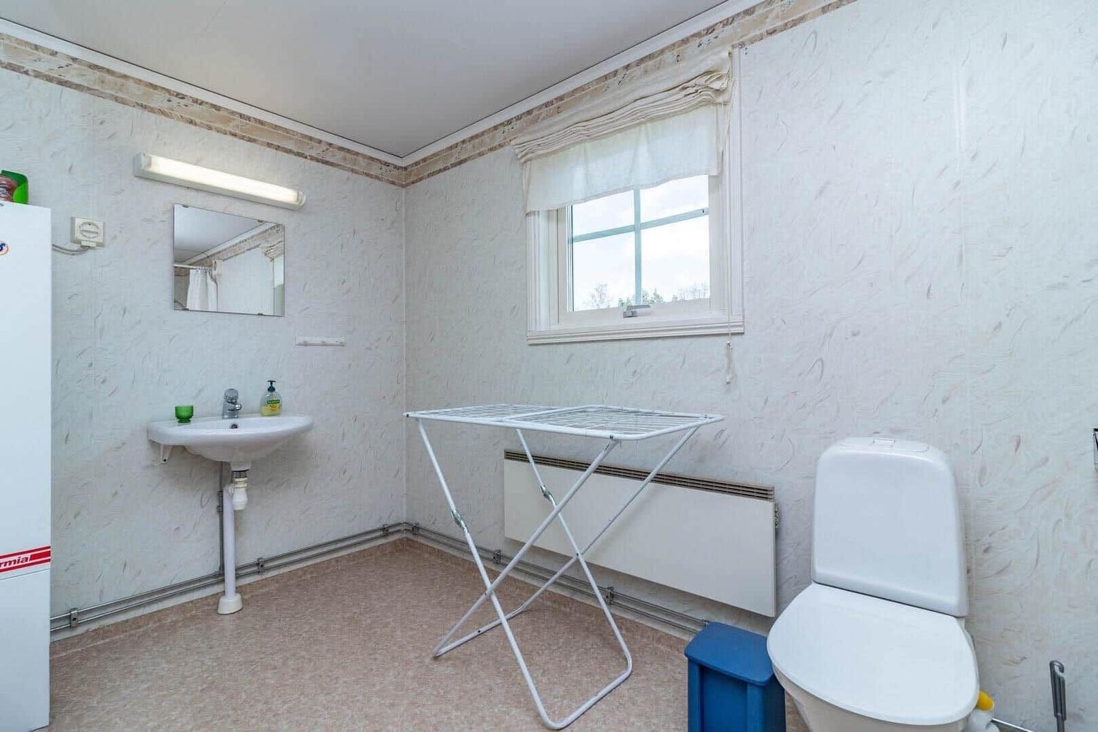Badezimmer mit Waschbecken, Toilette und Wäscheständer. Fenster mit Vorhang. Wandverkleidung und Bodenfliesen.