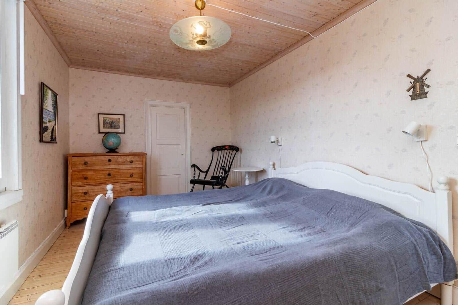 Schlafzimmer mit weißem Bett, Holzdecke und Schrank. Wanduhr und Stuhl sind sichtbar.