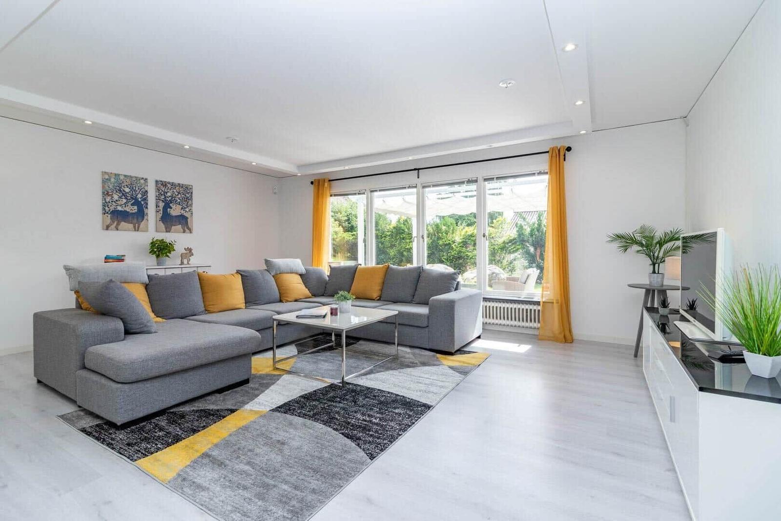 Wohnzimmer mit grauem Sofa, gelben Kissen und großem Fenster. Teppich und Wohnmöbel sind modern.