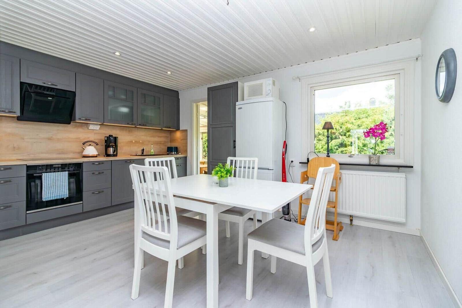 Küche mit Essbereich, weiße Tischstühle, graue Schränke und Fenster mit Blumen.