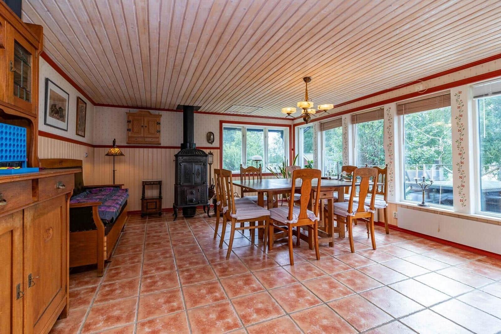Esszimmer mit Holztisch, Stühlen und Kachelboden. Fenster zeigen Garten. Ofen und Schränke sichtbar.
