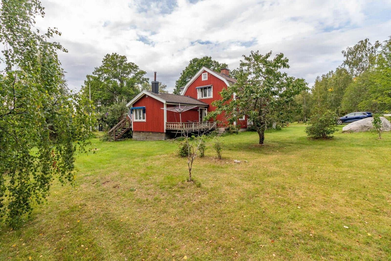 Rotes Holzhaus mit Terrasse, umgeben von Gras und Bäumen. Ein Auto steht auf dem Grundstück.
