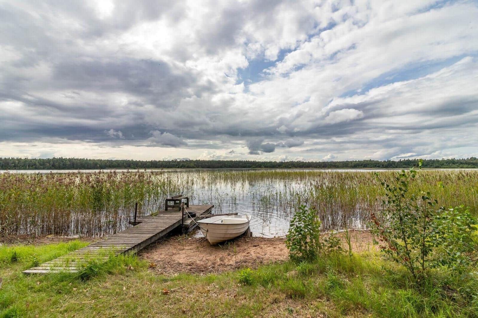 Ein weißes Boot liegt am Ufer eines Sees mit einem Holzsteg. Im Hintergrund sind Bäume und Wolken zu sehen.