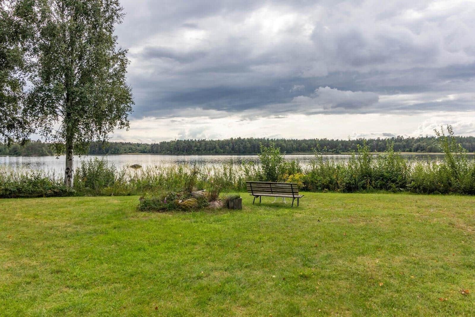 Ein Holzstuhl steht auf einer grünen Wiese am Ufer eines Sees unter bewölktem Himmel.