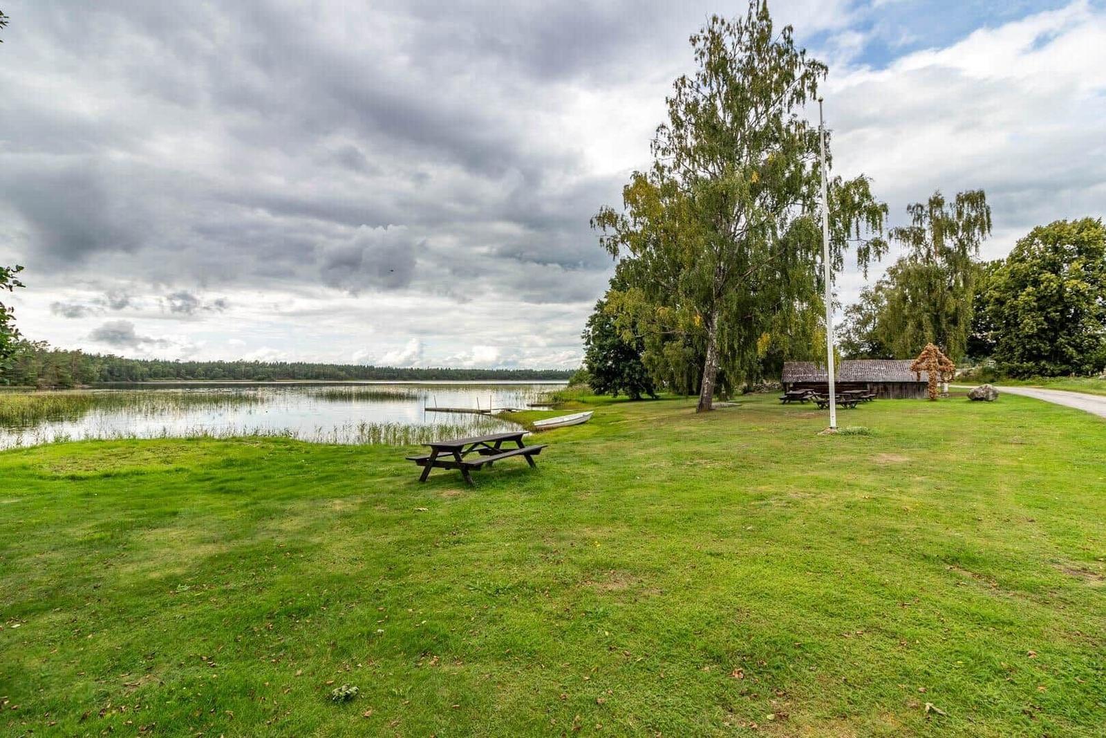 Grüner Rasenbereich am See mit Picknicktischen und Boot. Hintergrund: Wald und bewölkter Himmel.
