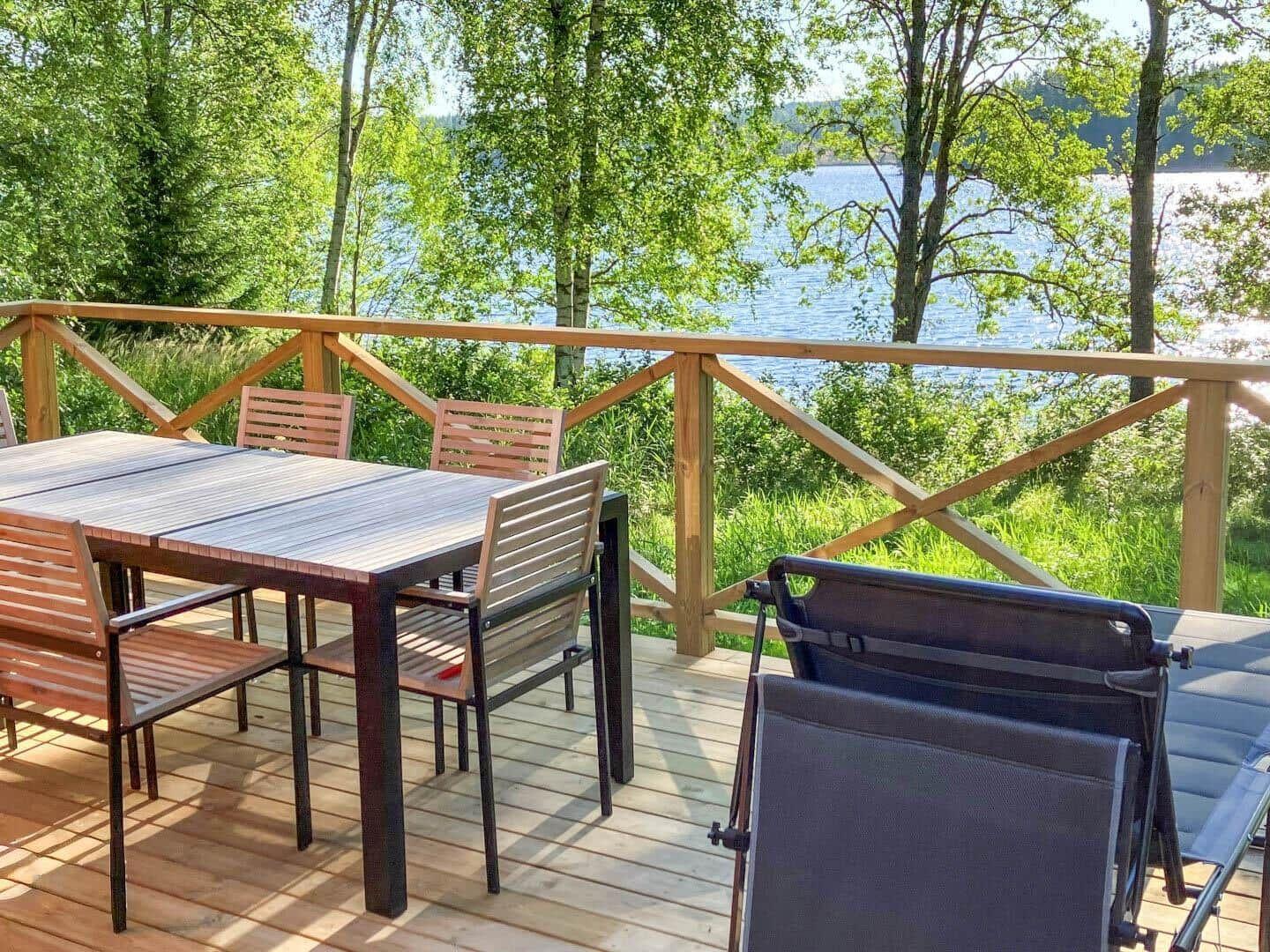 Holzterrasse mit Tisch, Stühlen und Liege, Blick auf See und Bäume.