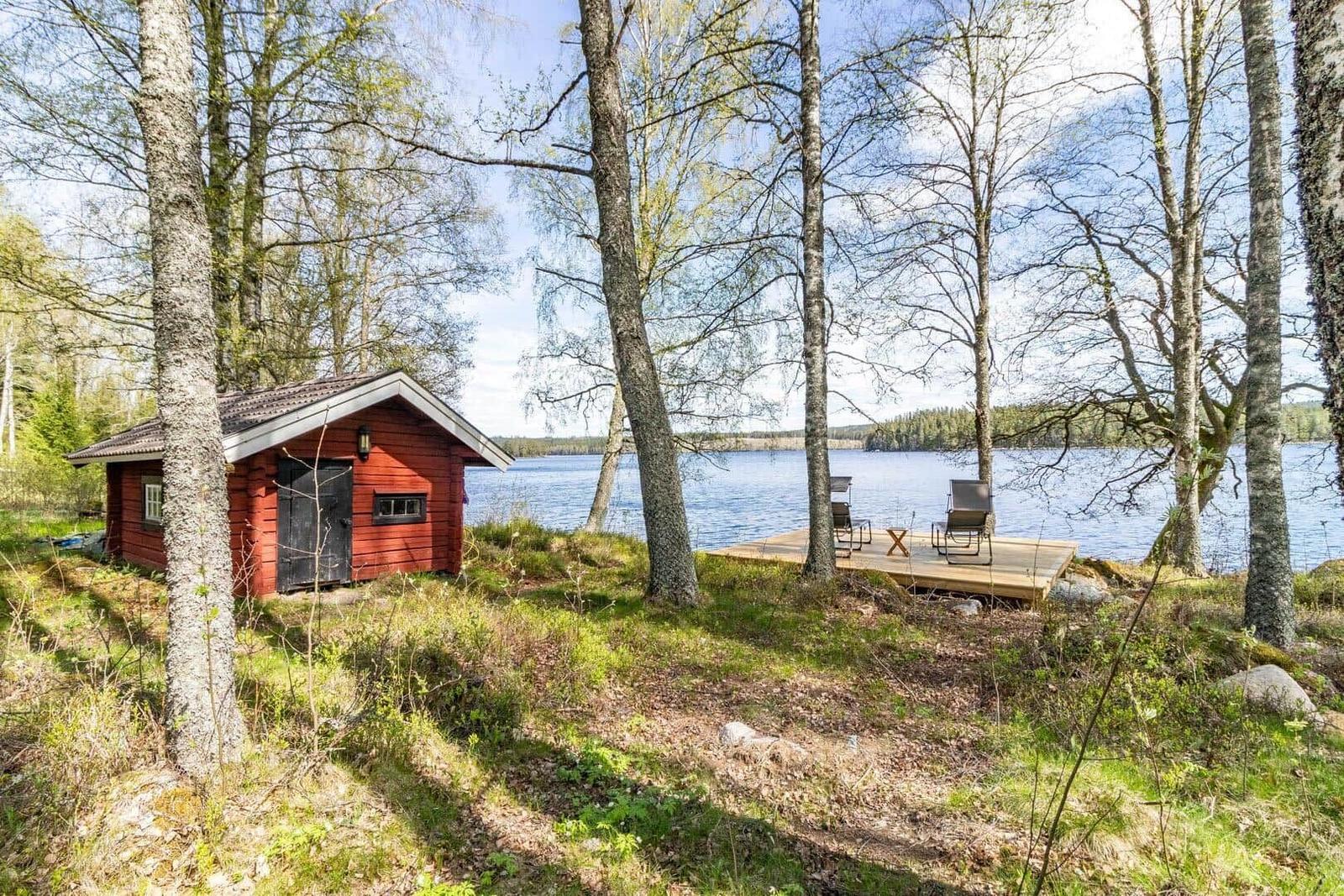 Rotes Holzhaus am See mit Terrasse und Stühlen zwischen Bäumen.
