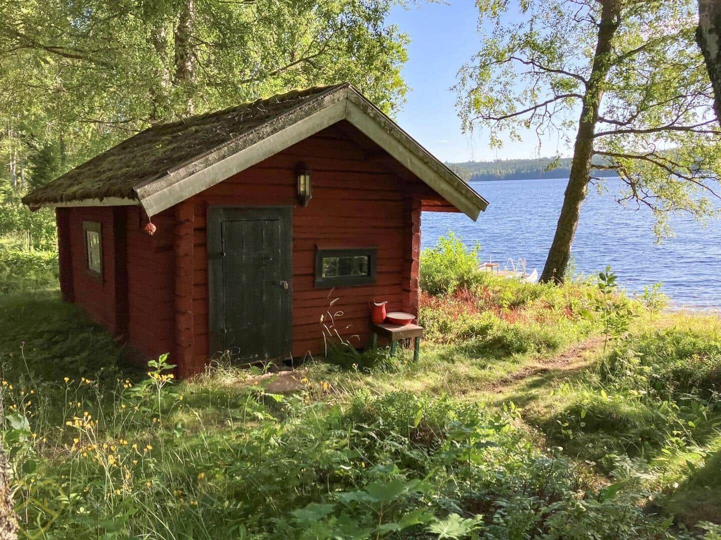 Rotes Holzhaus am See mit grüner Umgebung und einem kleinen Tisch vor der Tür.