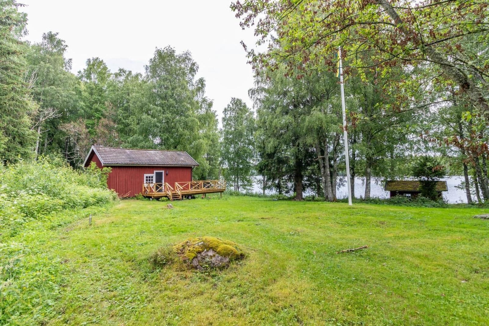 Rotes Holzhaus mit Terrasse, umgeben von Gras und Bäumen am See.