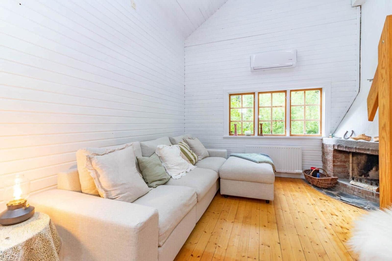 Wohnzimmer mit Sofa, Kamin und Holzboden. Fenster mit Blick auf Grün.