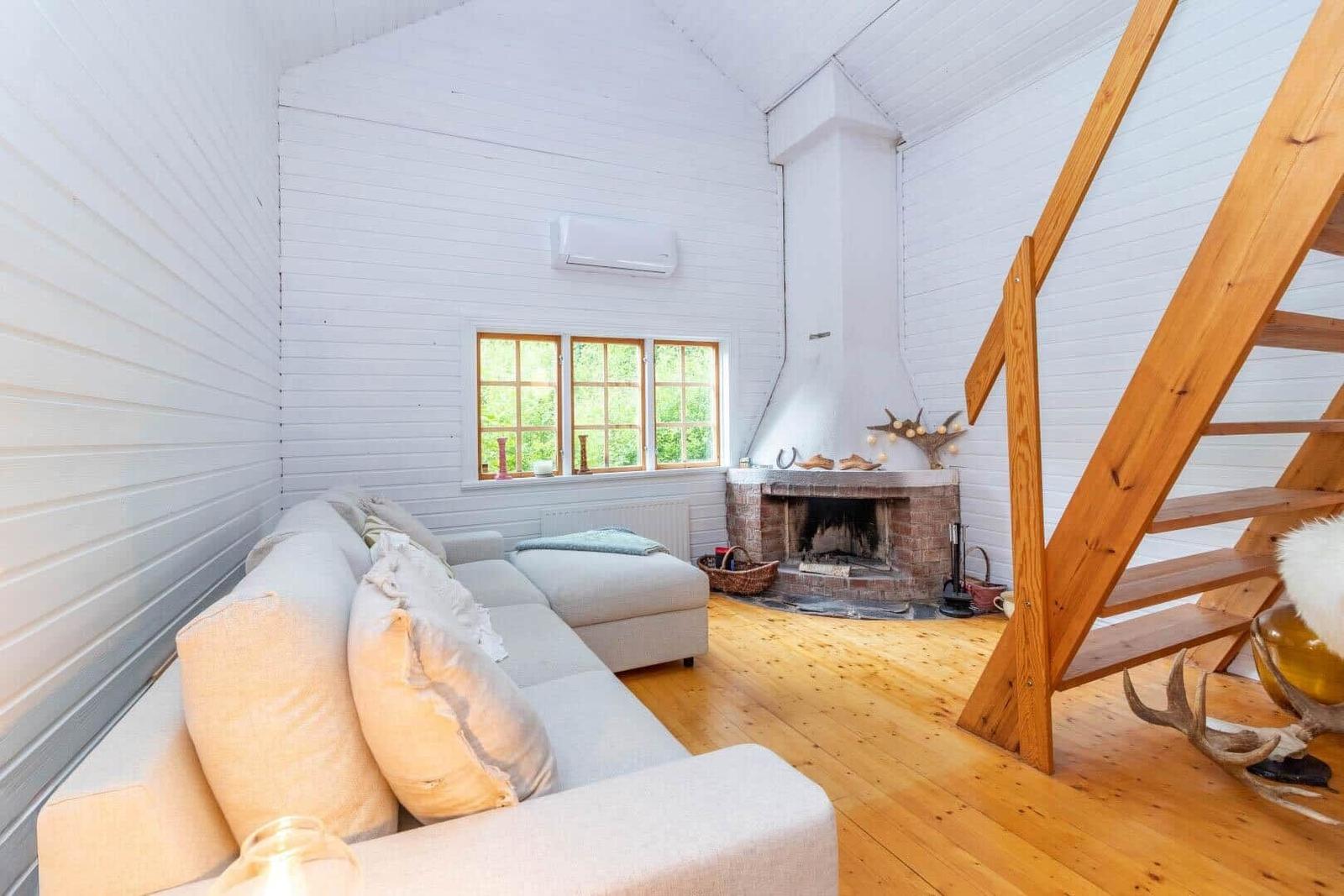 Wohnraum mit Sofa, Kamin und Holztreppe. Fenster mit Blick auf Grün.