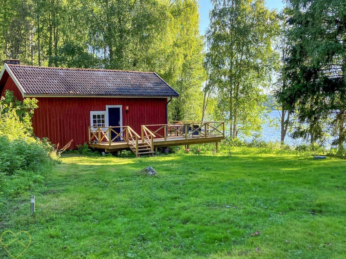Rotes Holzhaus mit Terrasse am See, umgeben von Bäumen und Gras.