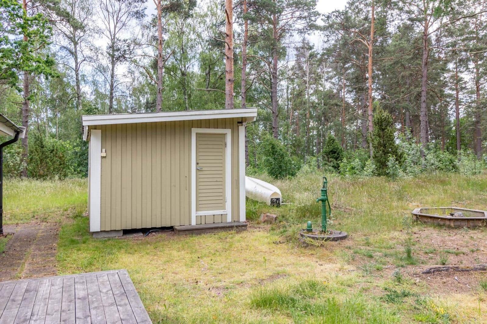 Wooden shed with door in the woods, next to green water pump and wooden terrace.