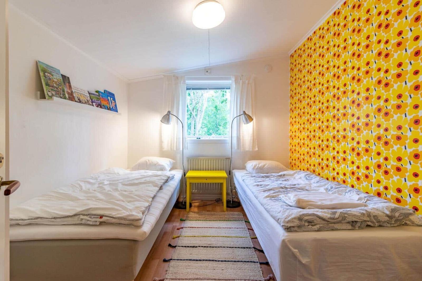 Two single beds, yellow table, window with green view, yellow-red patterned wall.