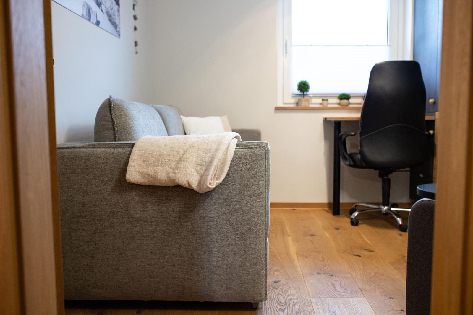 Ein grauer Sofa mit Decke, Schreibtisch und Stuhl im Raum.