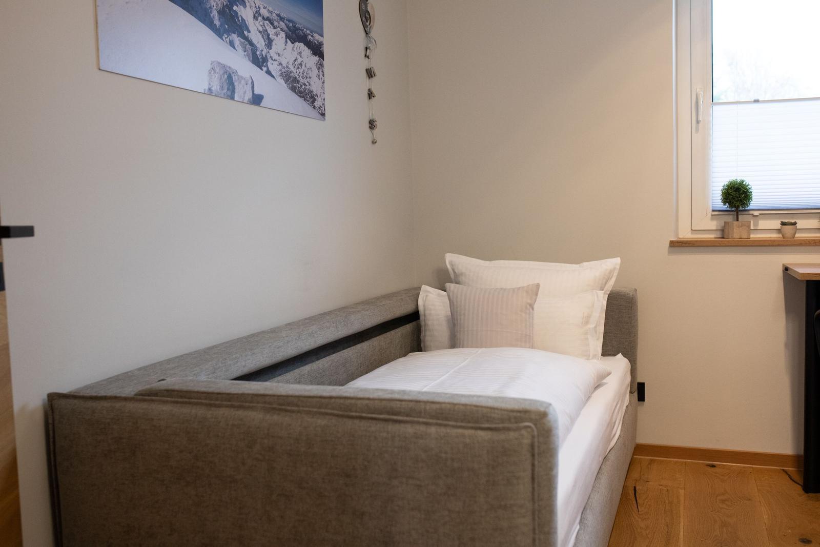 Ein Bett mit weißer Bettwäsche und Kissen. Fenster mit Jalousie und Pflanzen.