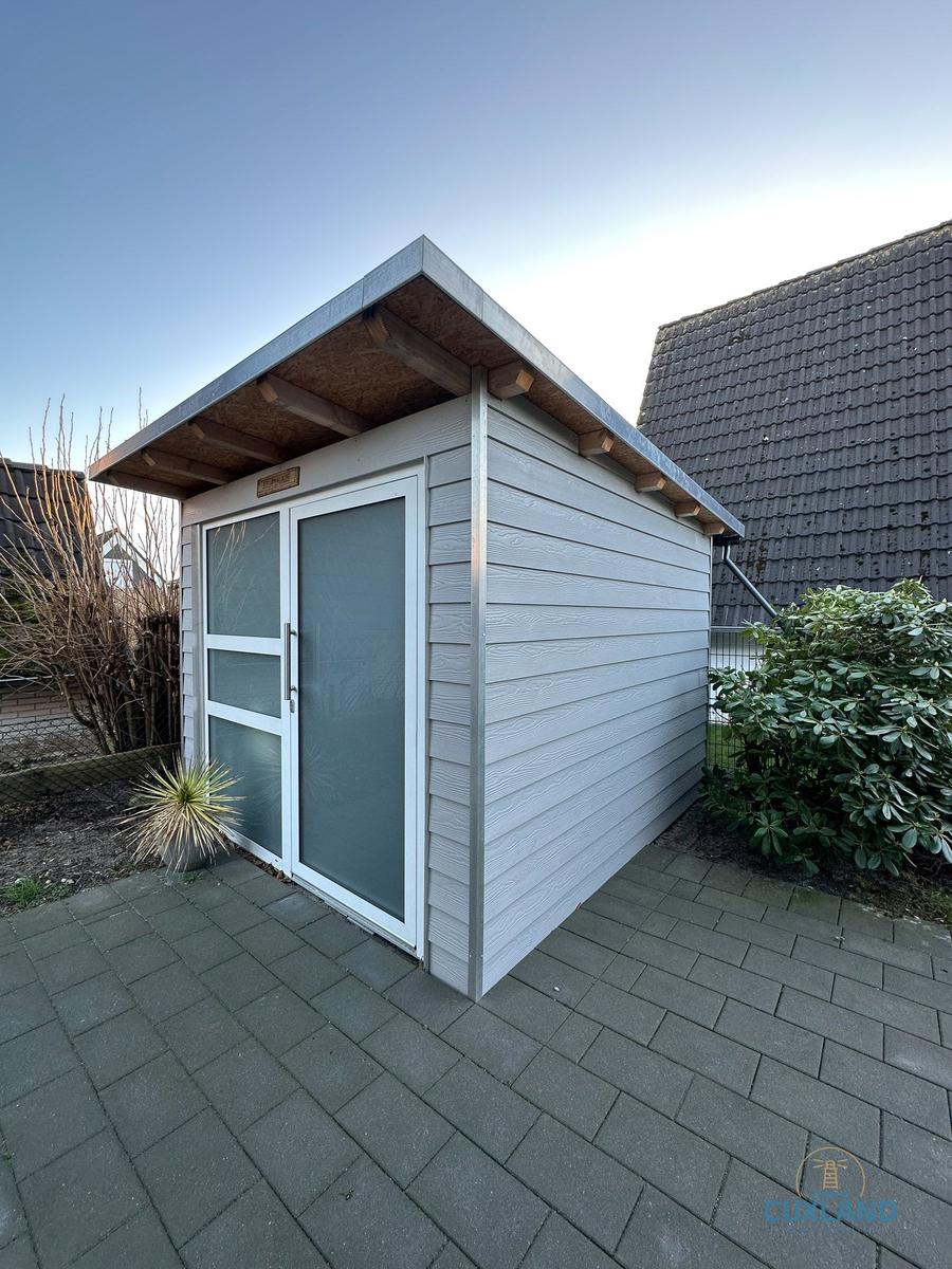 Ein kleines, graues Gartenhaus mit Glasfront und Dachvorsprung auf gepflastertem Boden.