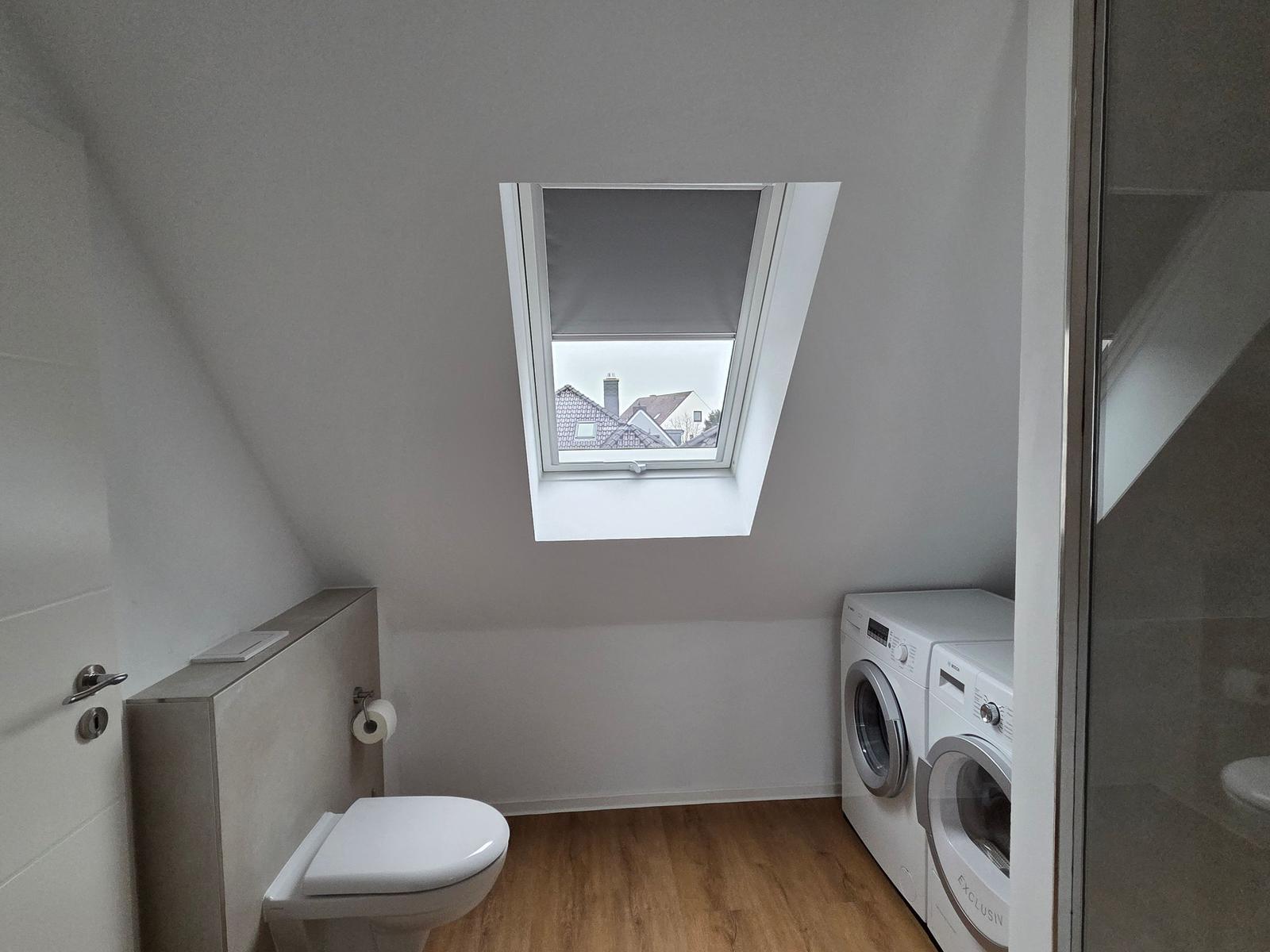 Duschbad mit Waschmaschine, Toilette und Dachfenster mit Blick auf Dächer.