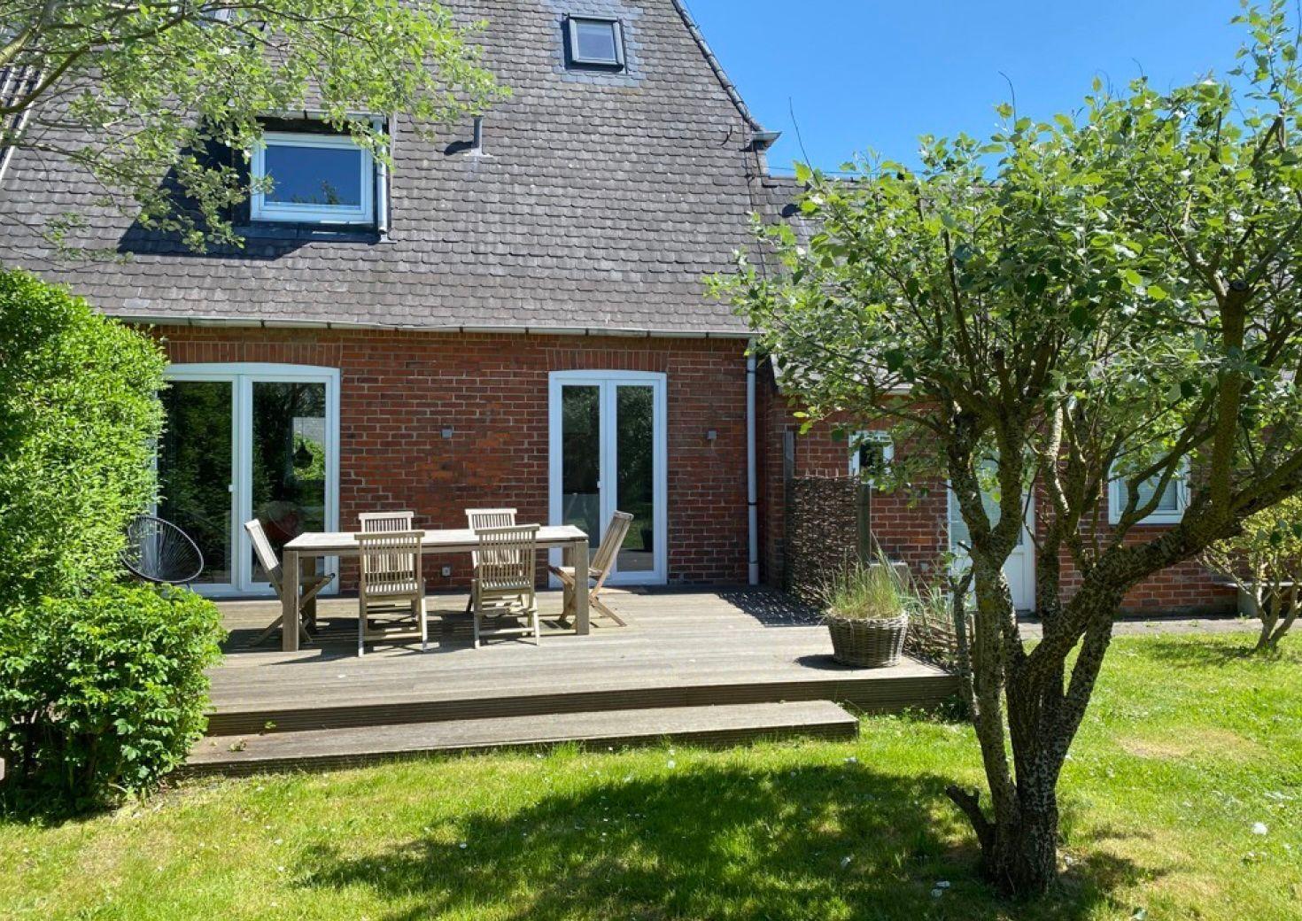 Haus mit Holzterrasse, Garten und Sitzgelegenheiten unter Bäumen.