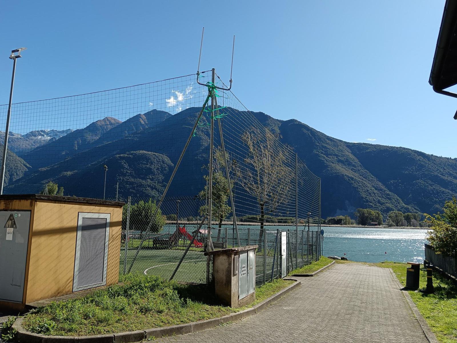Pfad entlang des Sees mit Sportplatz und Bergen im Hintergrund.