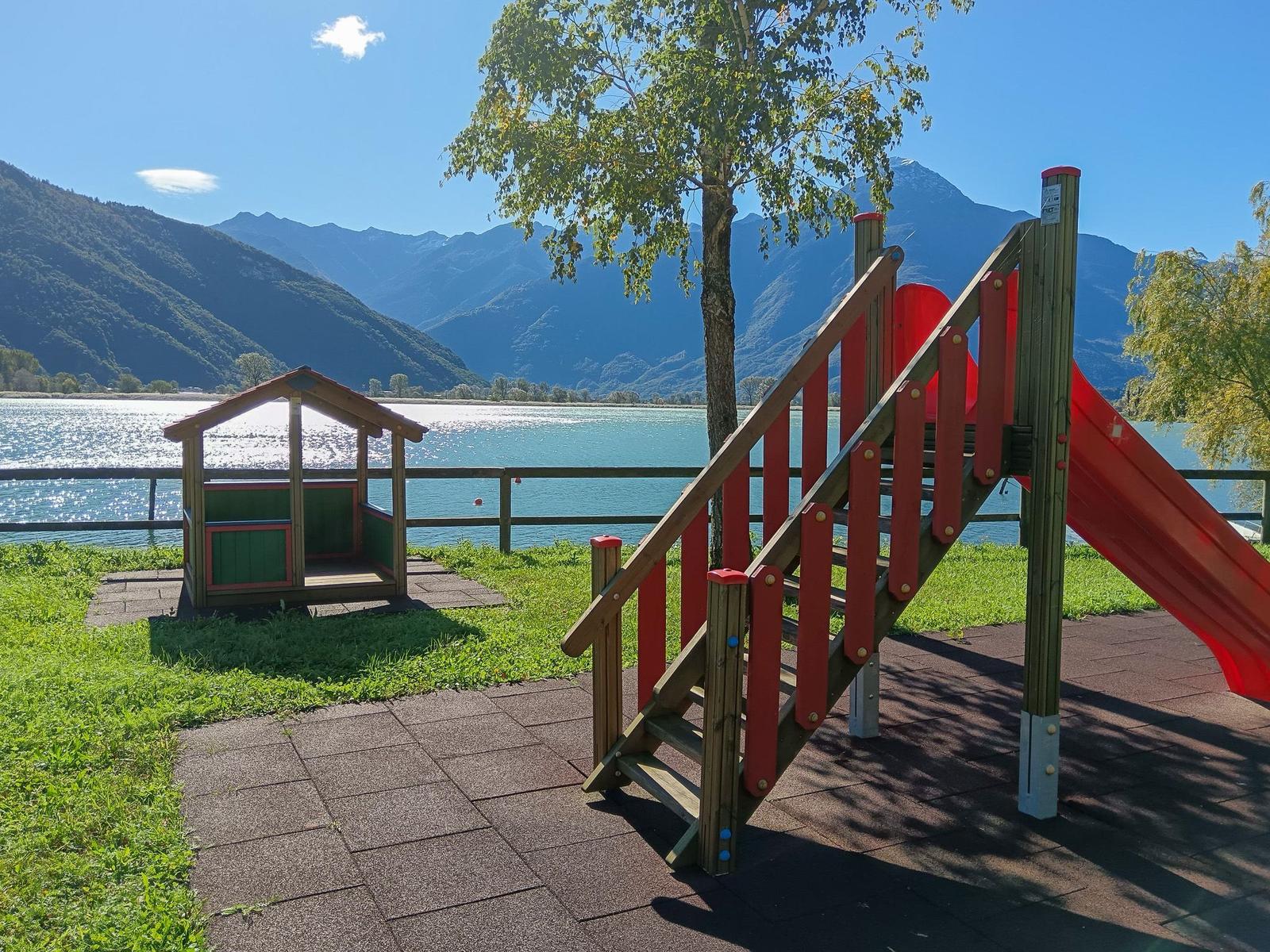 Kinderbereich mit Rutsche und Hütchen am See mit Bergen im Hintergrund.