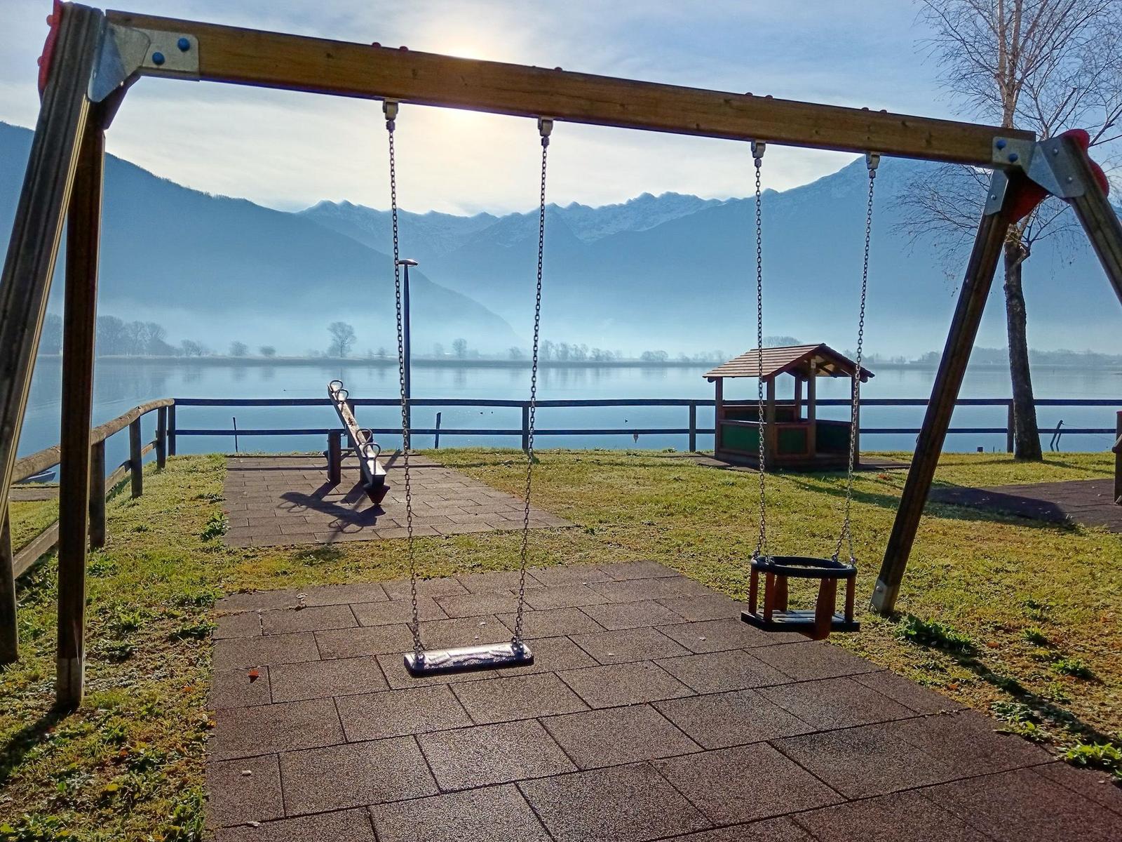 Kinderspielplatz mit Schaukel und Rutsche am See mit Bergen im Hintergrund.