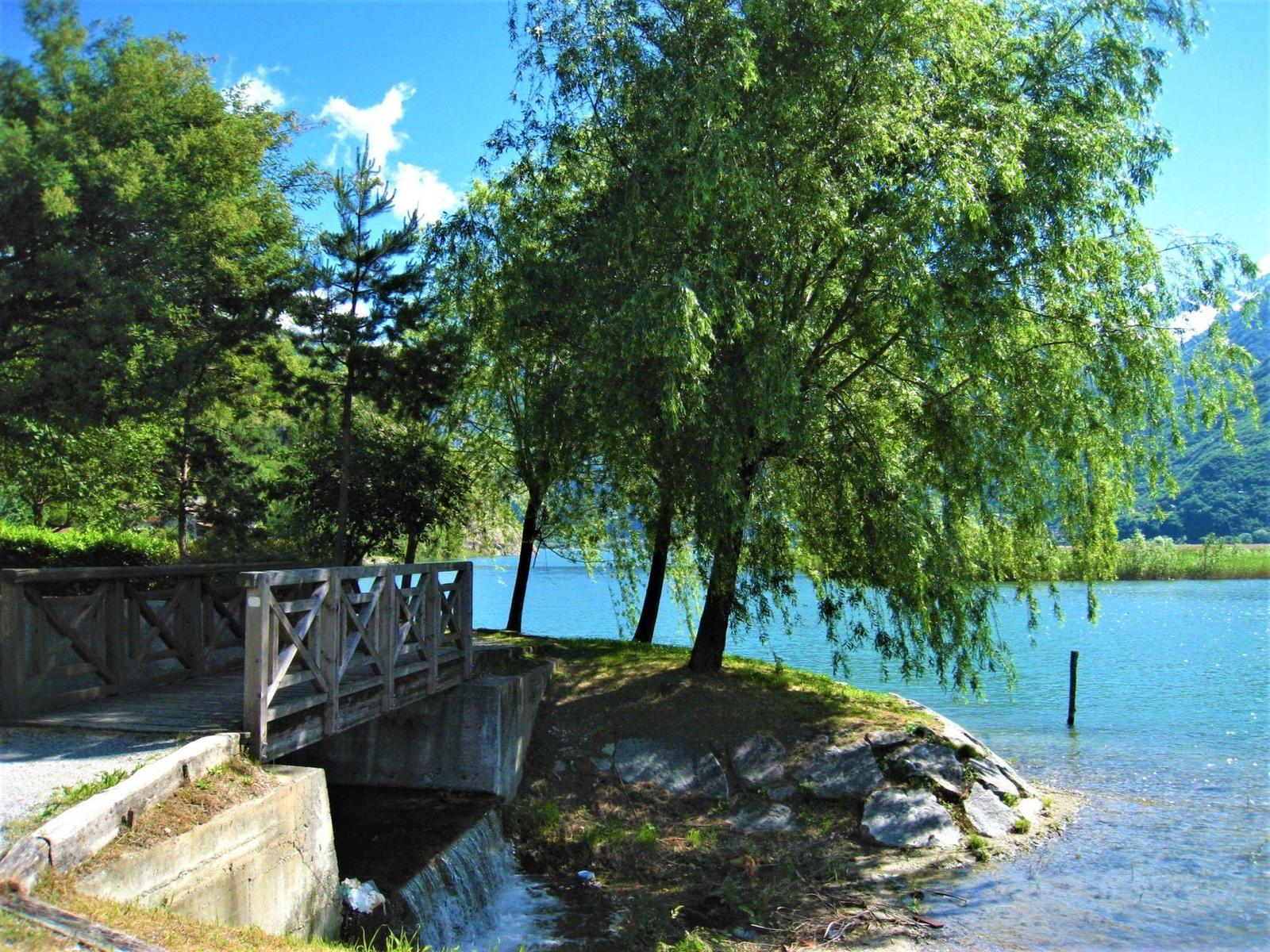 Holzbrücke über einen kleinen Wasserlauf am Seeufer mit Bäumen und Bergen im Hintergrund.