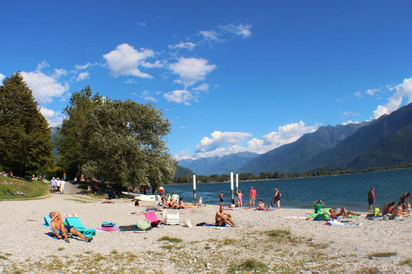 Strand mit Steinen, Menschen, Bäumen und Bergen im Hintergrund.