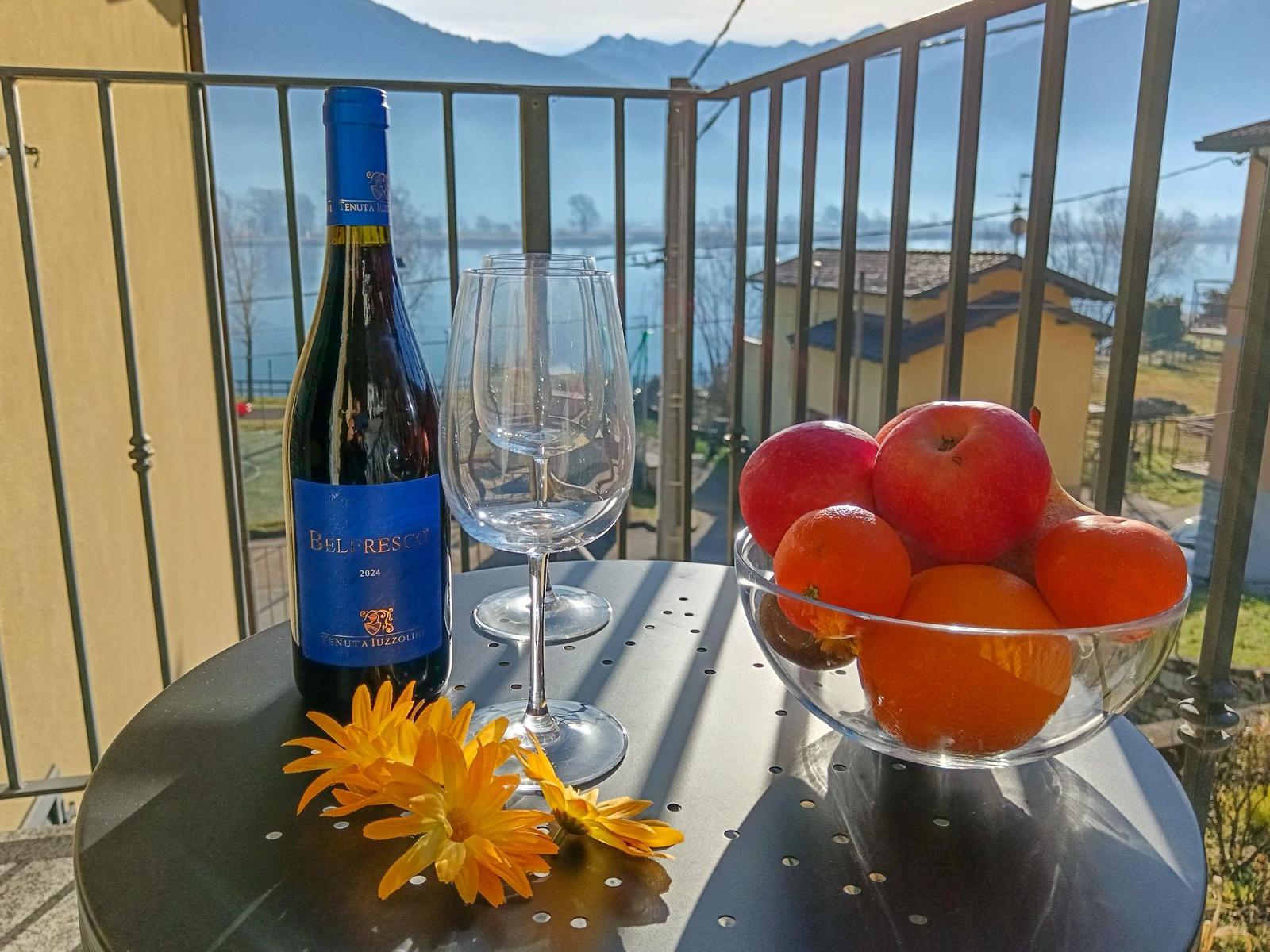 Auf der Terrasse steht ein Tisch mit Wein, Gläsern, Obst und Blumen. Im Hintergrund ist ein See und Berge zu sehen.