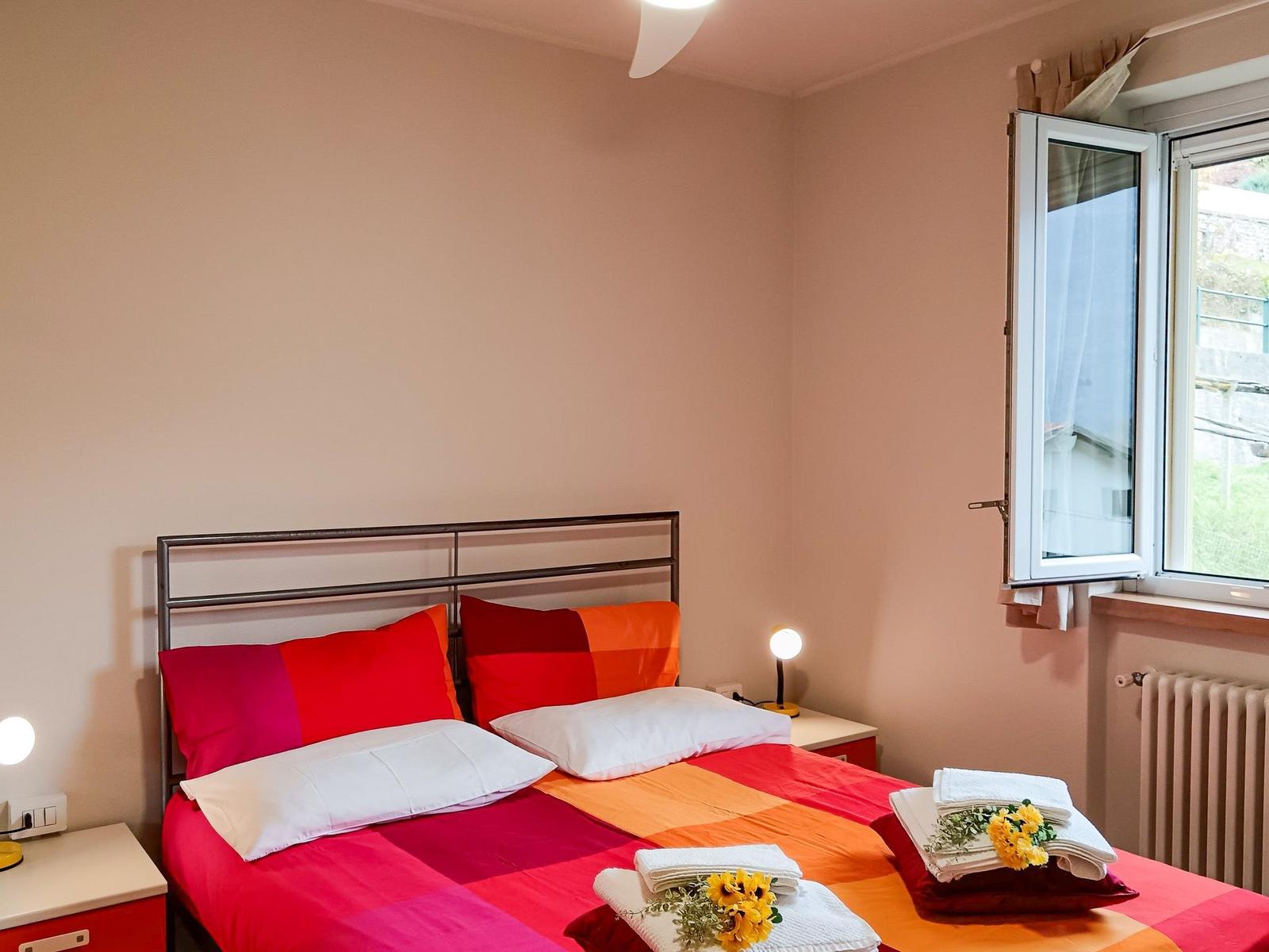Schlafzimmer mit Bett, Nachttischen und Fenster. Bett mit bunten Kissen und Handtüchern.