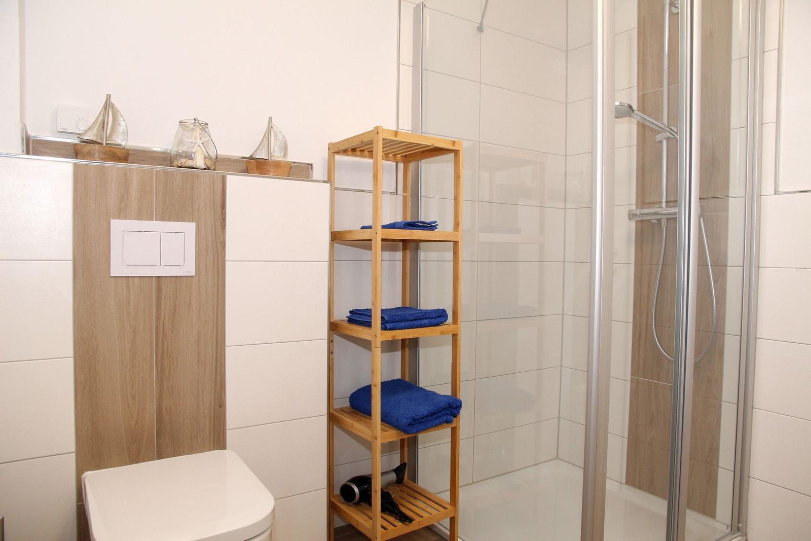 Badezimmer mit Dusche, Holzregal und Toilette. Blaue Handtücher und Deko auf Regal.