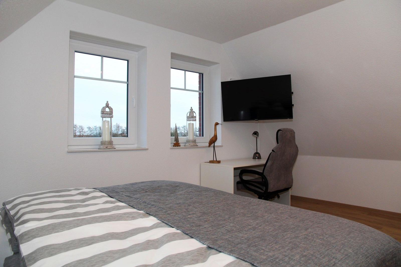 Schlafzimmer mit Bett, Schreibtisch, TV und zwei Fenstern.
