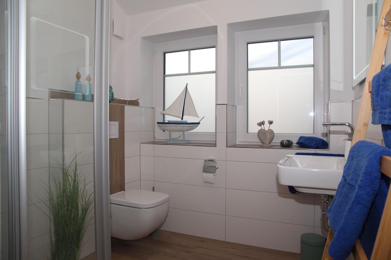 Badezimmer mit Dusche, Toilette, Waschbecken und Fenstern. Dekoration mit Segelboot und Handtuch.