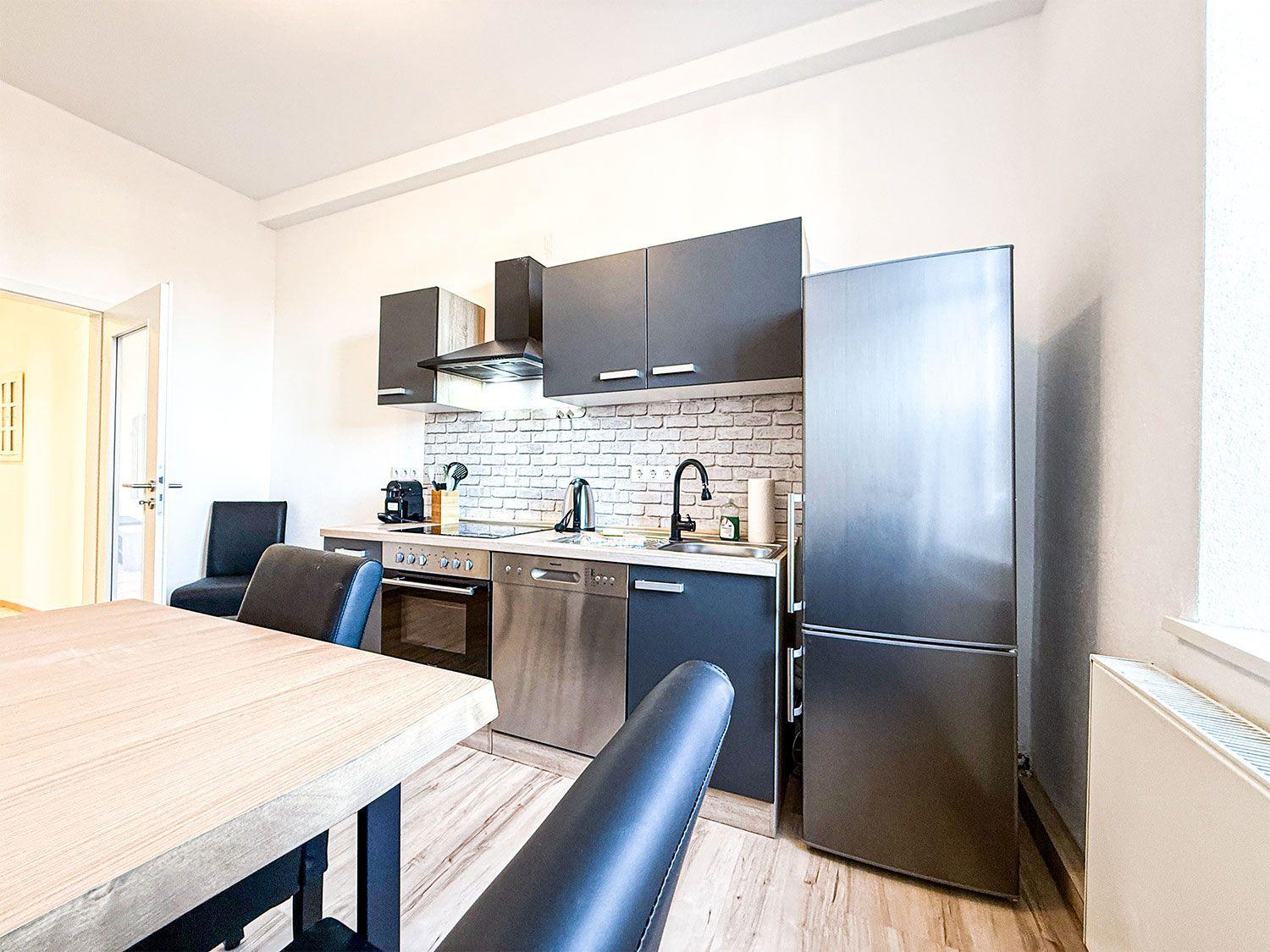 Küche mit Edelstahlgeräten, Holztisch und schwarzen Stühlen. Hintergrund: Backsteinwand und Kühlschrank.