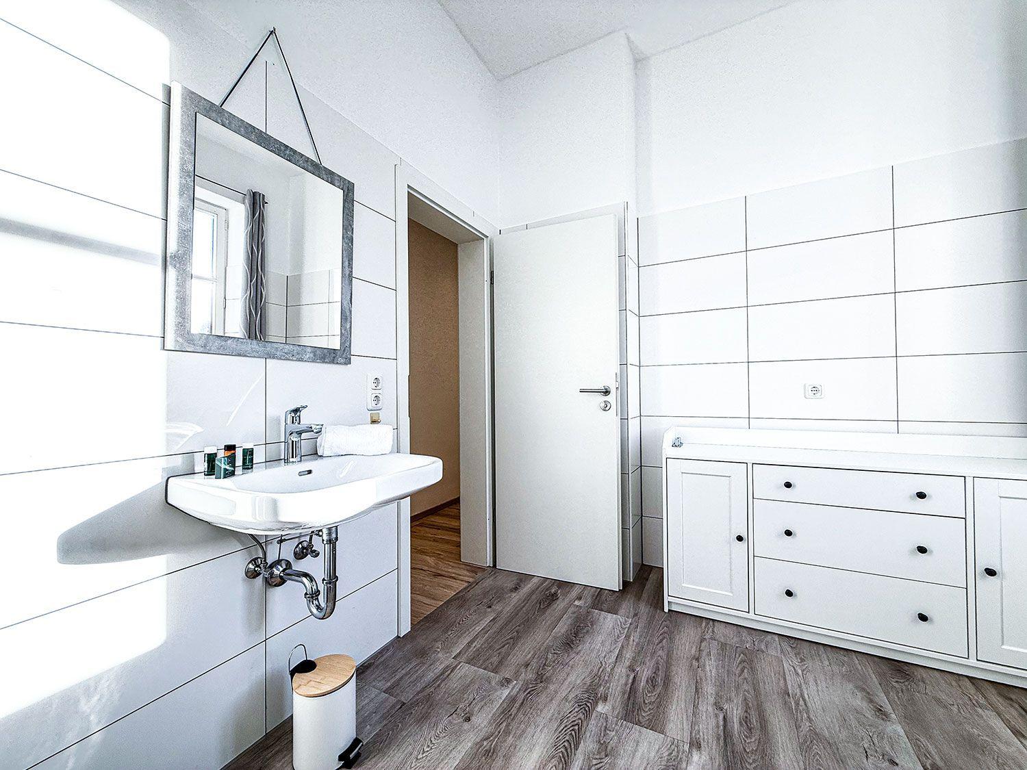 Badezimmer mit Waschbecken, Spiegel und Schrank. Holzdielenboden und weiße Fliesen.