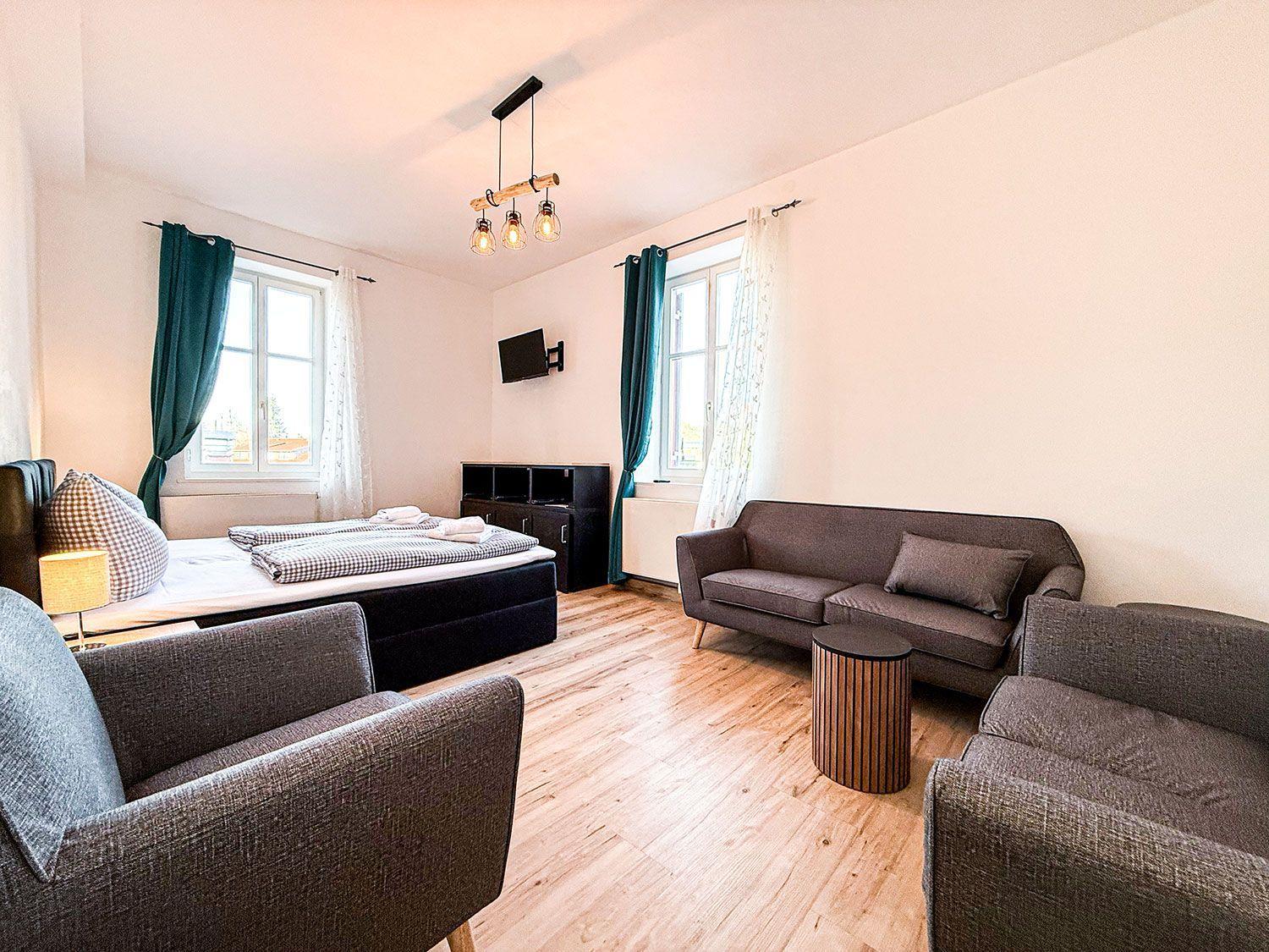 Zimmer mit Bett, Sofa, Stühlen und TV. Holzboden und Fenster mit Vorhängen.