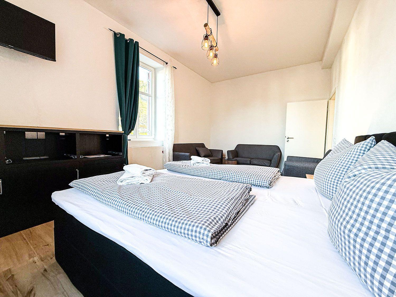 Zimmer mit Doppelbett, Sofa, Fenster und TV. Deckenleuchte und Holzboden.