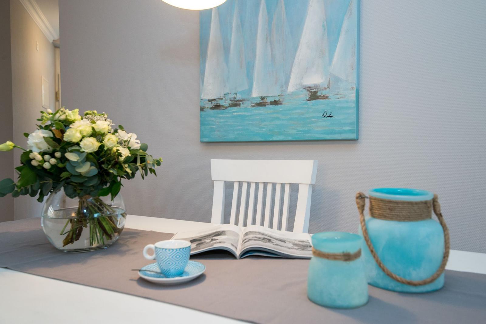 Tisch mit Blumen, Tasse, Buch und blauen Vasen. Wandbild mit Segelbooten.