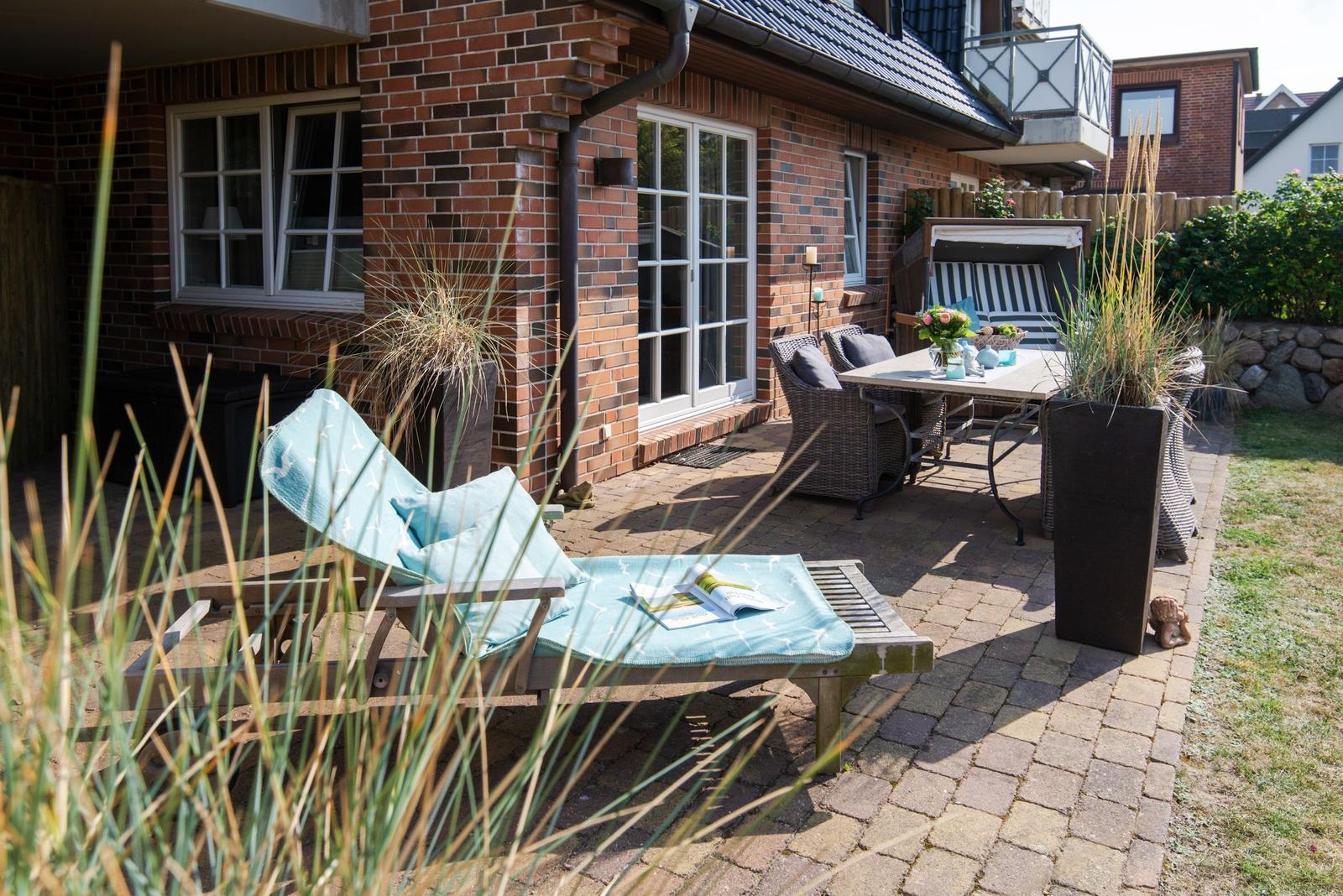 Terrasse mit Liegestuhl, Tisch und Stühlen. Hintergrund: Backsteinhaus mit Fenstern.