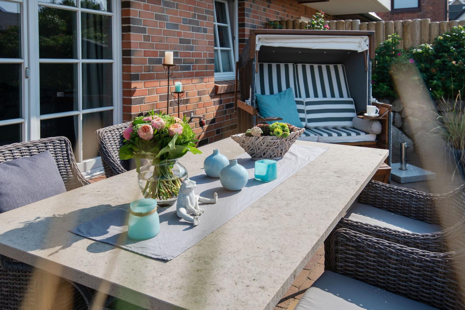 Terrasse mit Tisch, Stühlen und Sonnenliege. Blumen, Vasen und Kerzen dekorieren den Außenbereich.