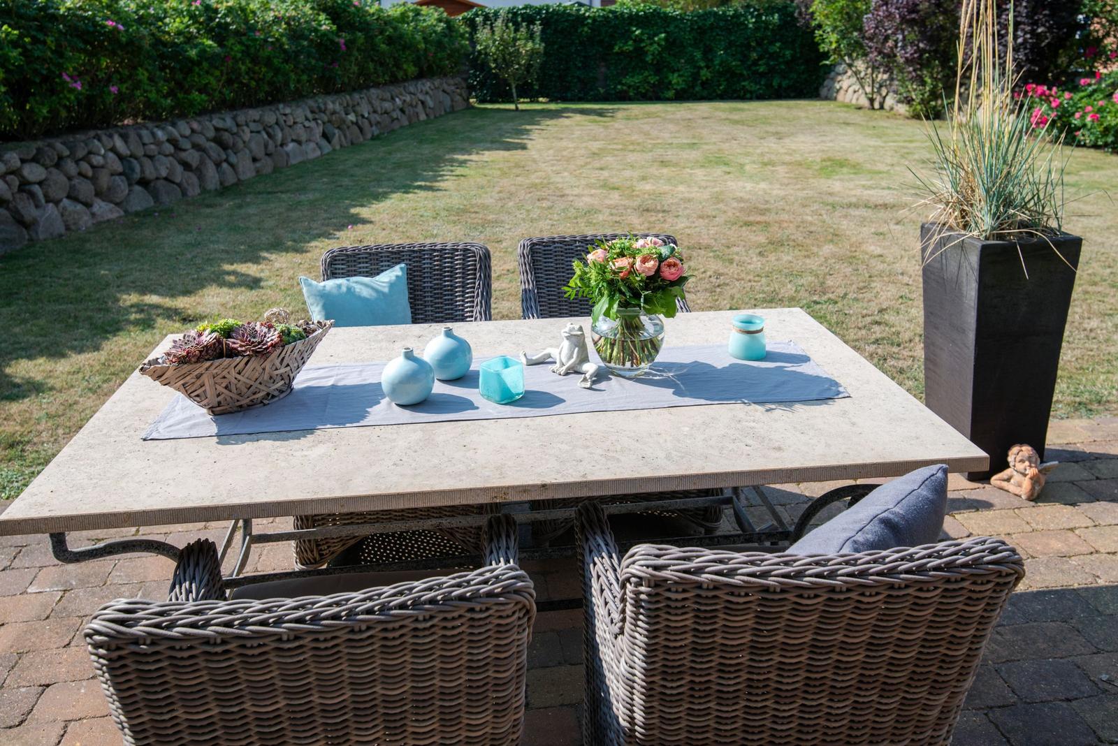 Garten-Terrasse mit gedecktem Tisch und Stühlen. Hintergrund: Rasen und Steinmauer.