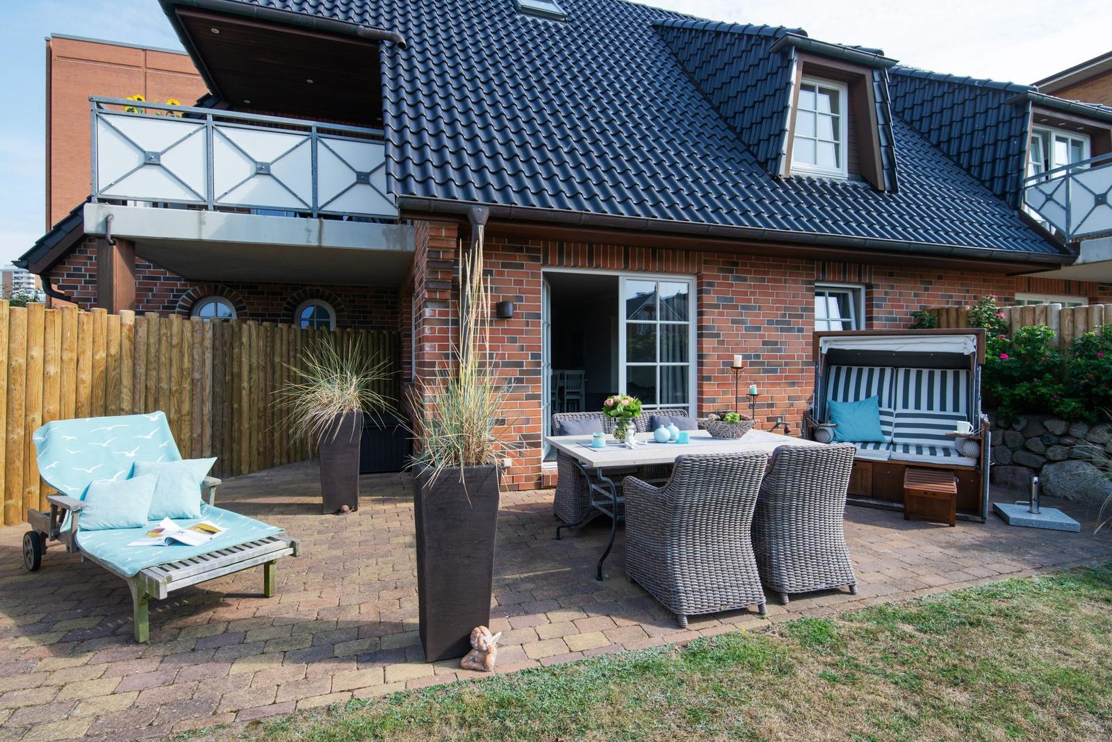 Hinterhof mit Terrasse, Tisch, Stühlen und Liege. Holzzaun und Garten.