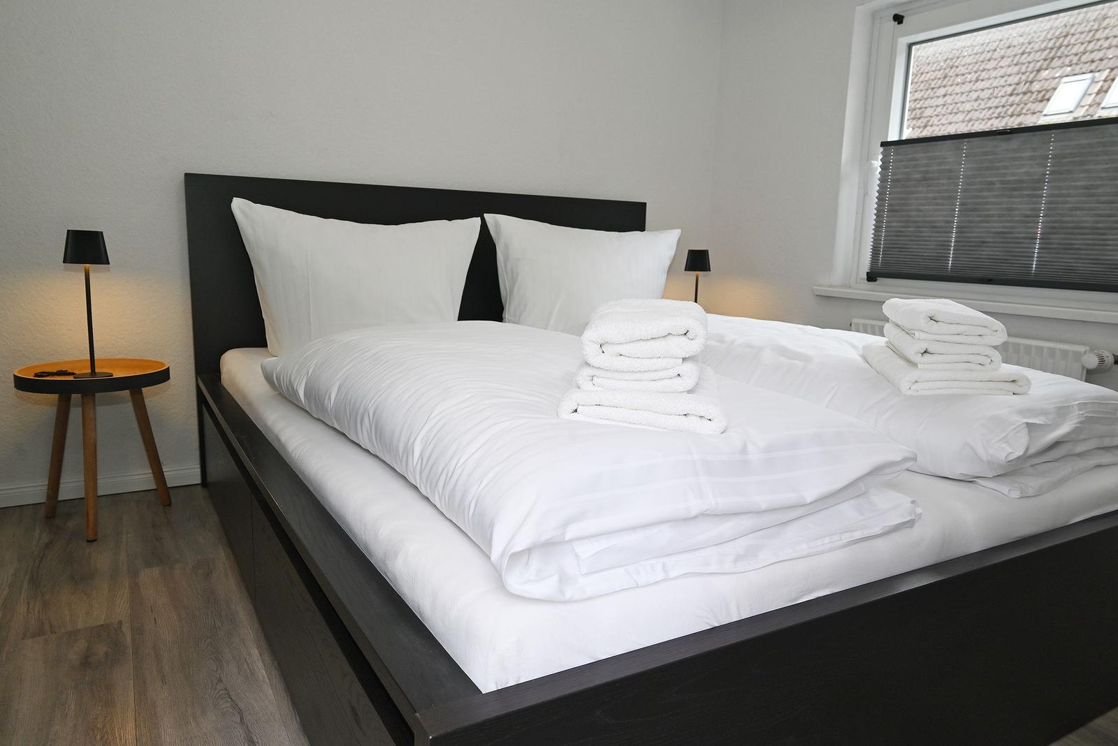 Ein Bett mit weißer Bettwäsche und gefalteten Handtüchern. Links steht ein Nachttisch mit Lampe.