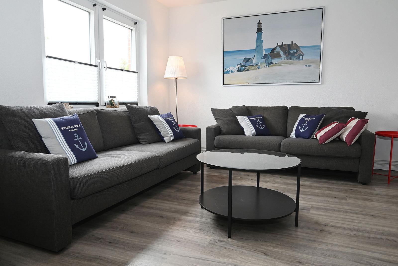 Zwei graue Sofas mit Kissen stehen um einen runden Tisch herum. An der Wand hängt ein Leuchtturm-Bild.