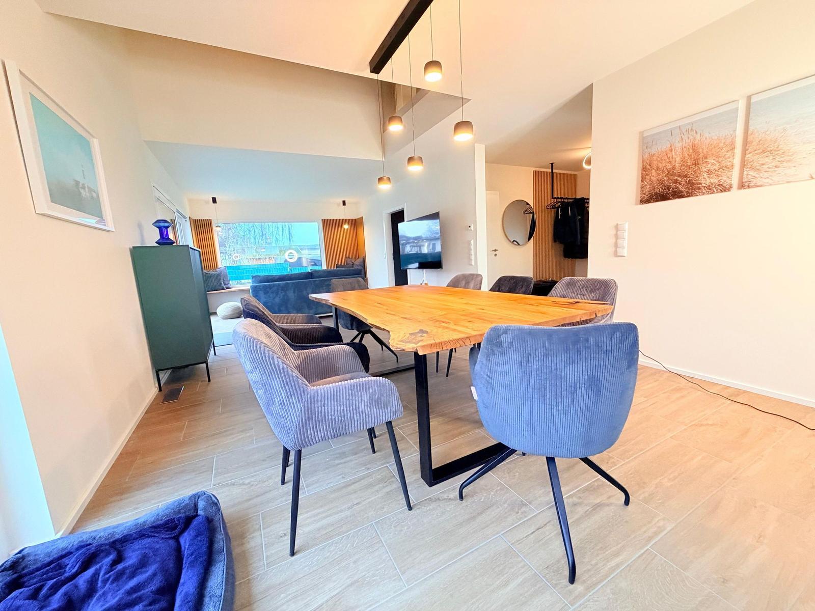 Essbereich mit Holztisch und blauen Stühlen. Im Hintergrund Wohnbereich mit Sofa und Fernseher.
